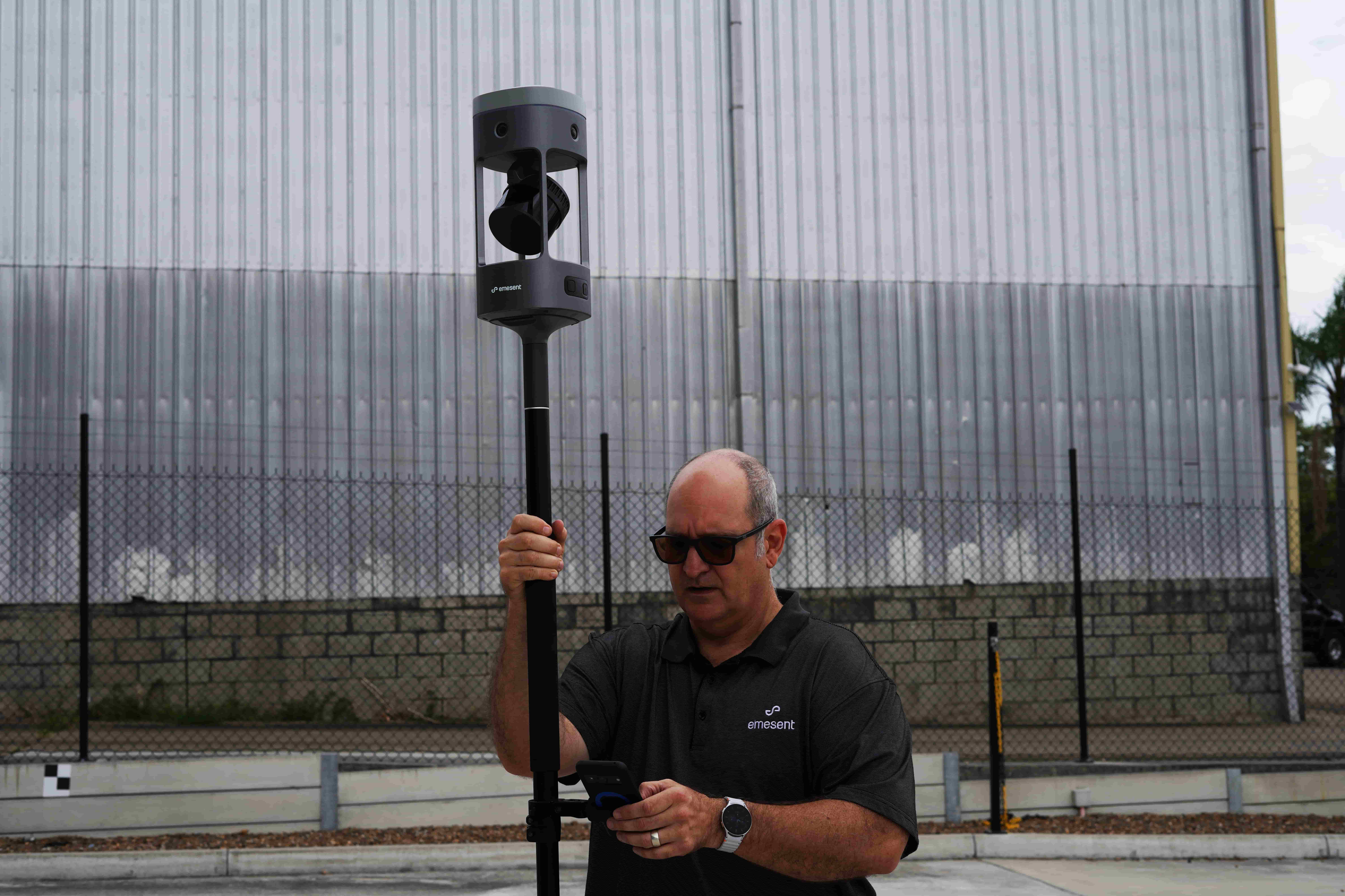 A man wearing sunglasses adjusts a surveying instrument mounted on a tall pole in an industrial outdoor setting.