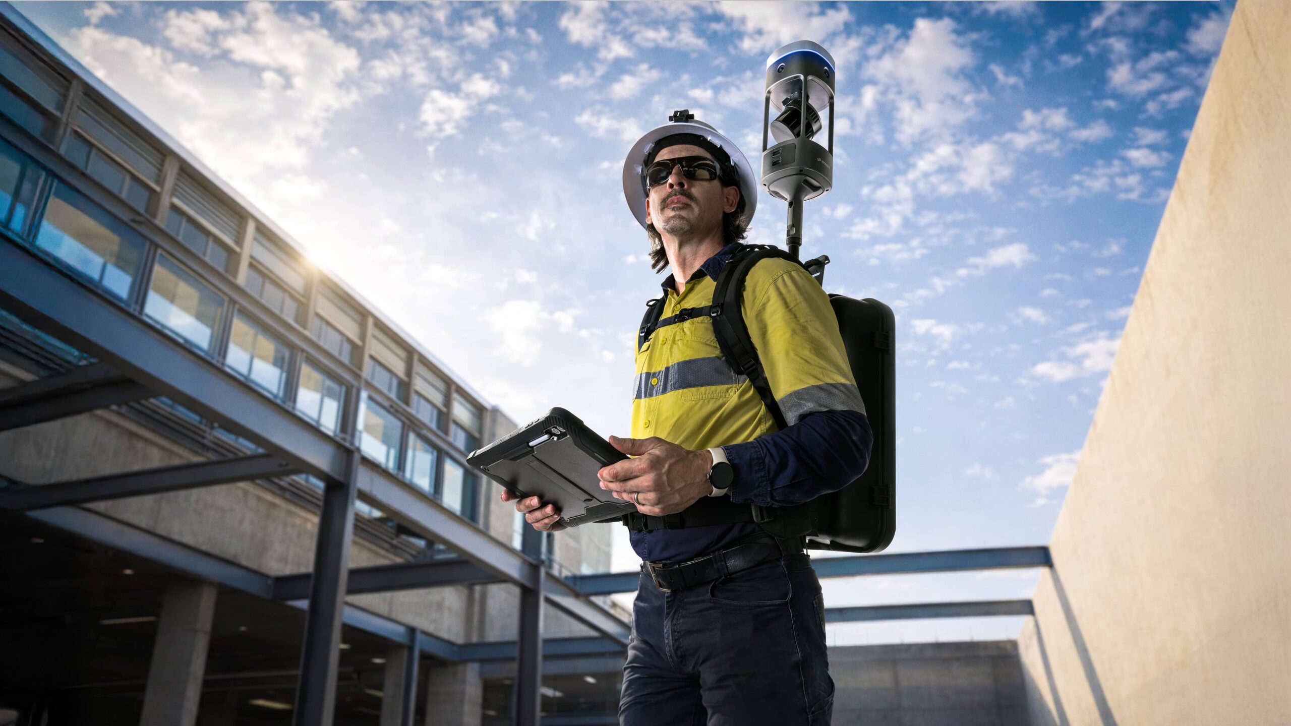 Drone operator in safety gear and helmet uses a tablet controller outdoors near a modern building.