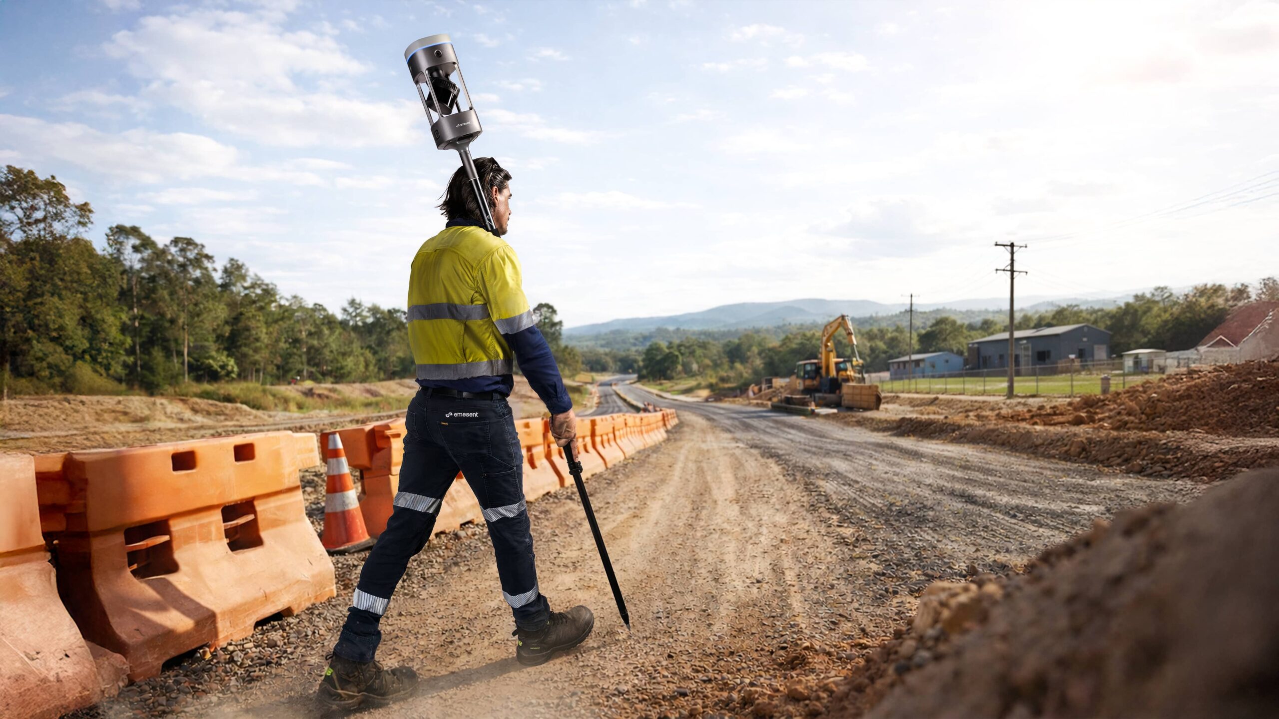 Construction worker in a high-visibility yellow vest walks along a dirt road with a surveying pole on a road construction site, orange barriers lining the path.