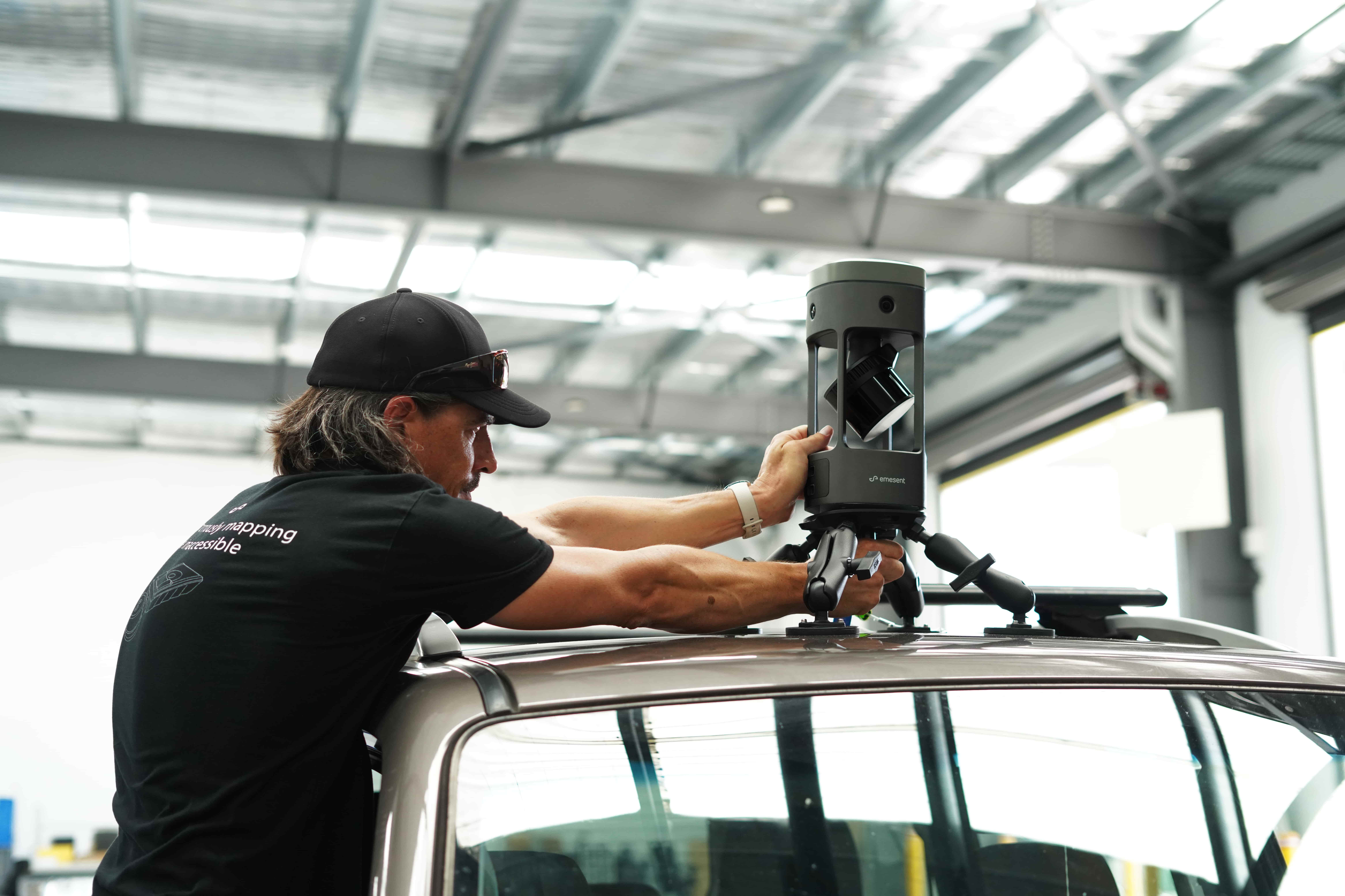 Technician in a cap mounts a car-mounted 3D laser scanner on the vehicle roof inside a workshop.