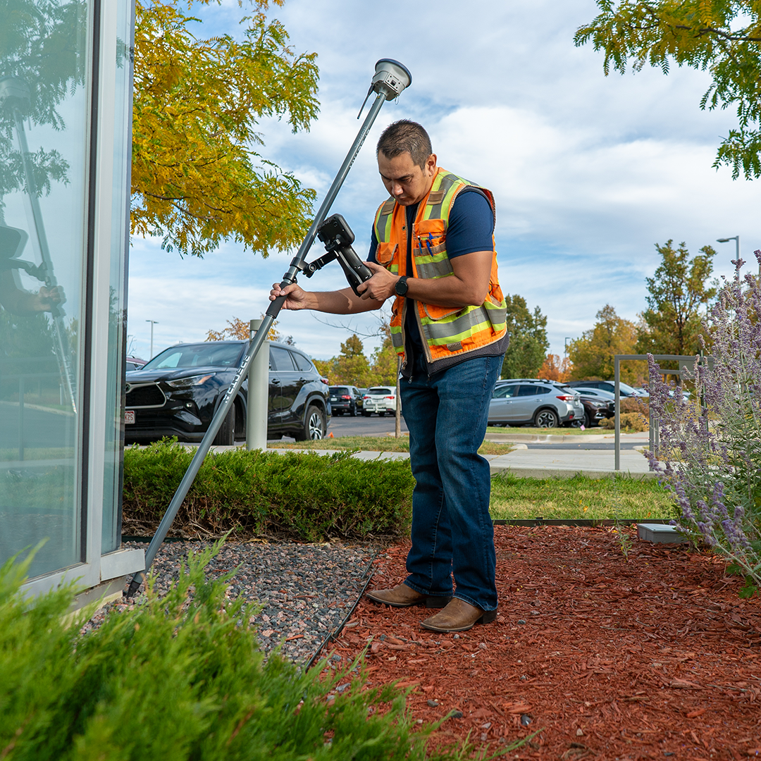 Construction worker in a safety vest and hard hat uses a handheld device beside a tripod-mounted surveying instrument outdoors.