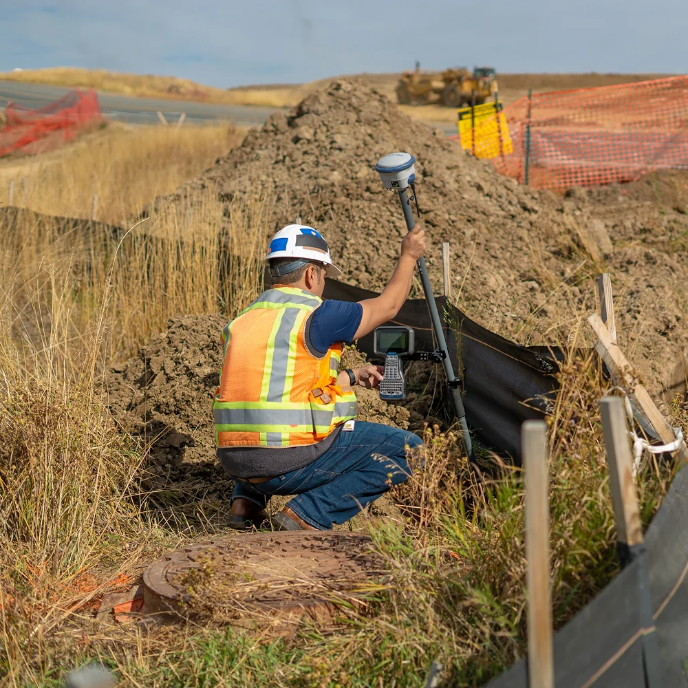 Construction worker in a safety vest and hard hat uses a surveying instrument at a dirt trench on a road construction site.