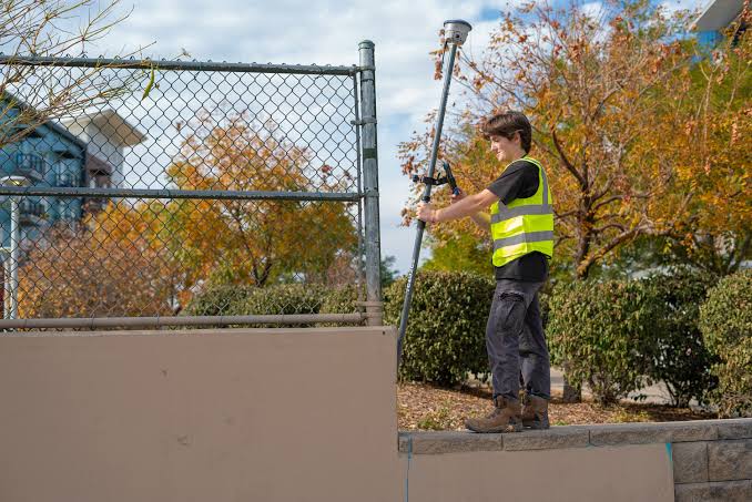 Worker in a neon safety vest uses a long tool to adjust a chain-link fence outdoors on a sunny autumn day.
