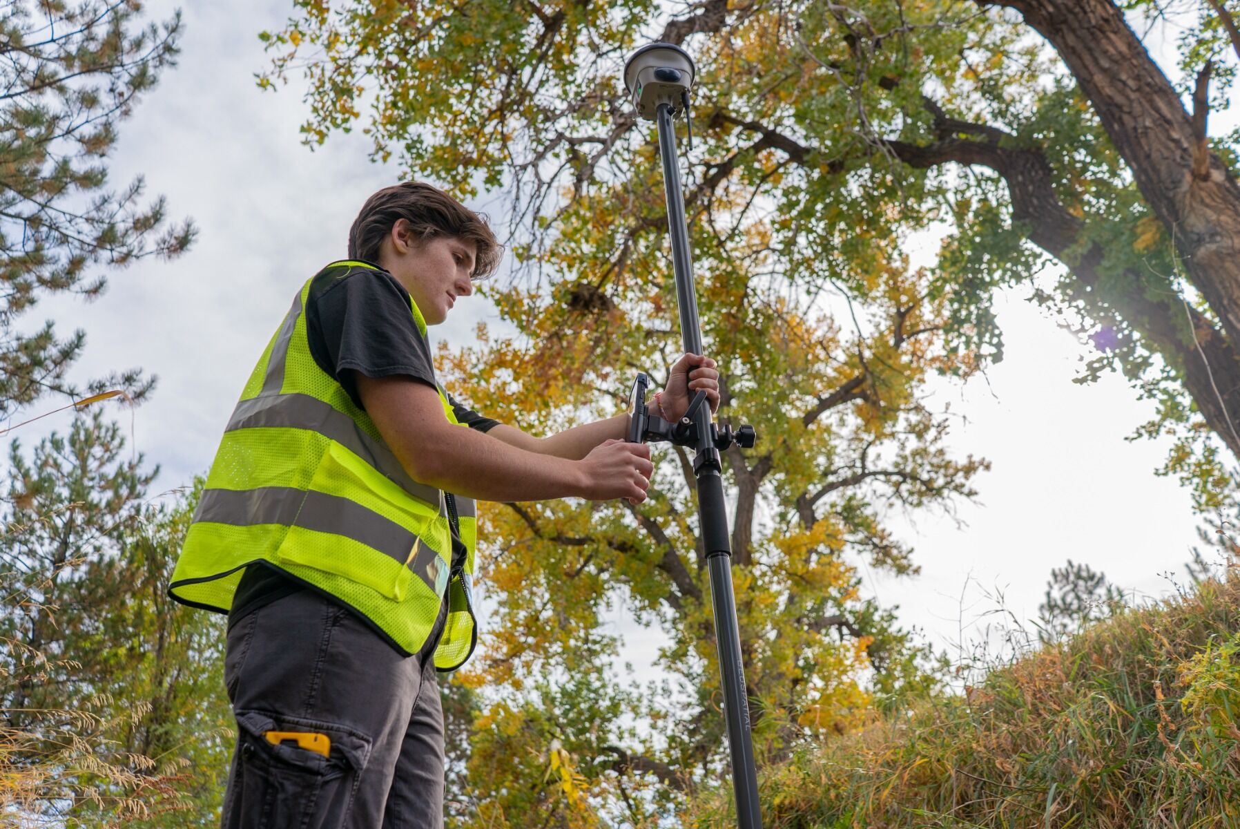 Technician in a neon safety vest adjusts a tripod-mounted surveying instrument outdoors among trees.
